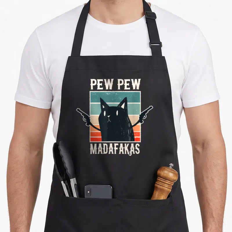 Madafakas cat apron - Black cat holding two guns with PEW PEW text above and MADAFAKAS below on a striped background — shown on a black cotton canvas apron against a white background