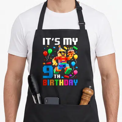 9th birthday building aprons - Block figure holding cake surrounded by balloons and IT'S MY 9TH BIRTHDAY text — shown on a black cotton canvas apron against a white background