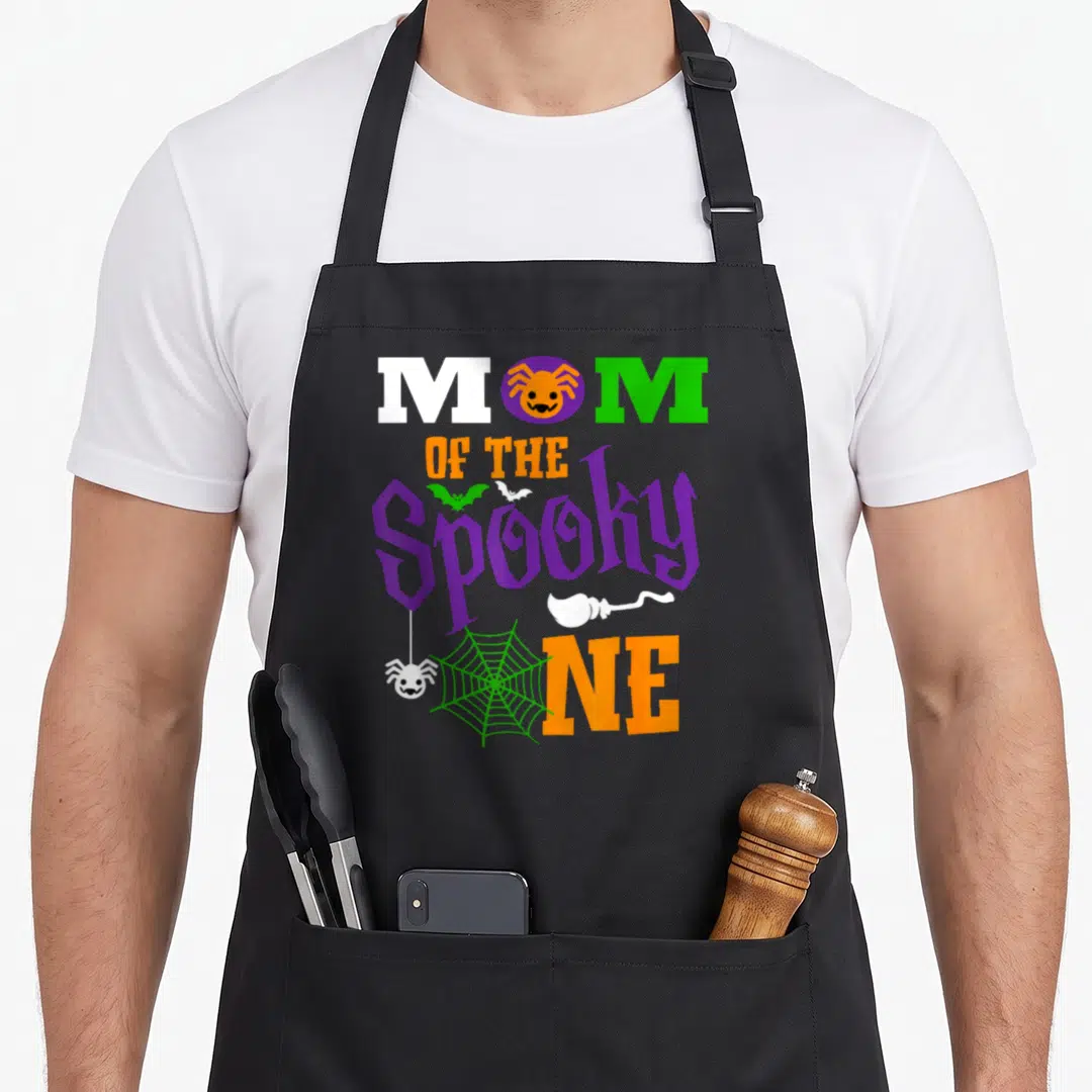 1st funny halloween apron - Stacked Mom of the Spooky One text with cartoon spiders bats and ghost in purple orange — shown on a black cotton canvas apron against a white background