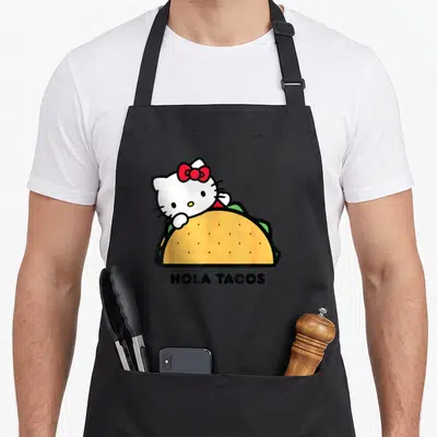 Hola taco apron - Hello Kitty peeking from behind yellow taco with HOLA TACOS text below — shown on a black cotton canvas apron against a white background