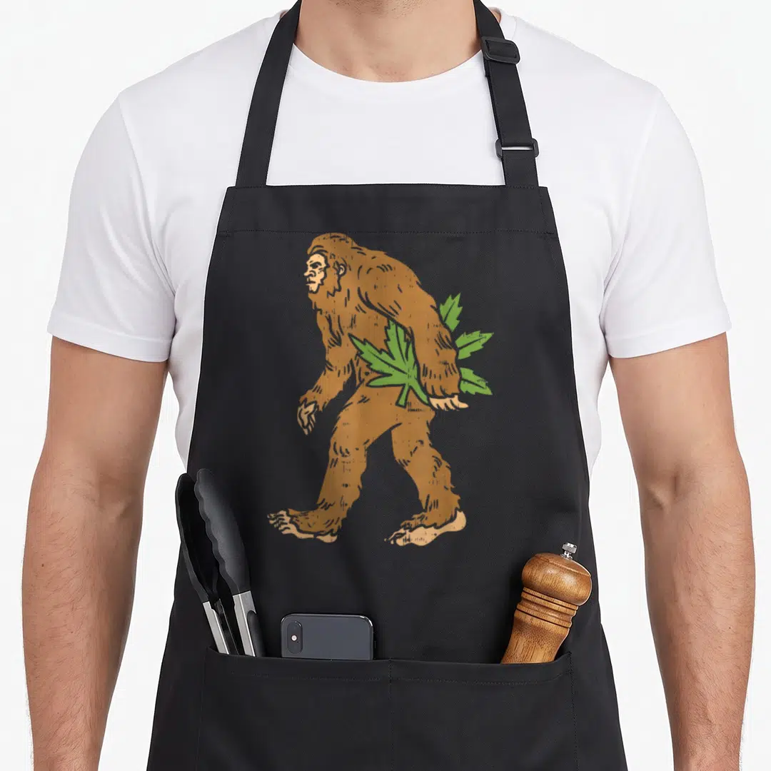 Boredkoalas bigfoot apron - Retro brown Bigfoot walking left with green leaves behind back on black apron — shown on a black cotton canvas apron against a white background