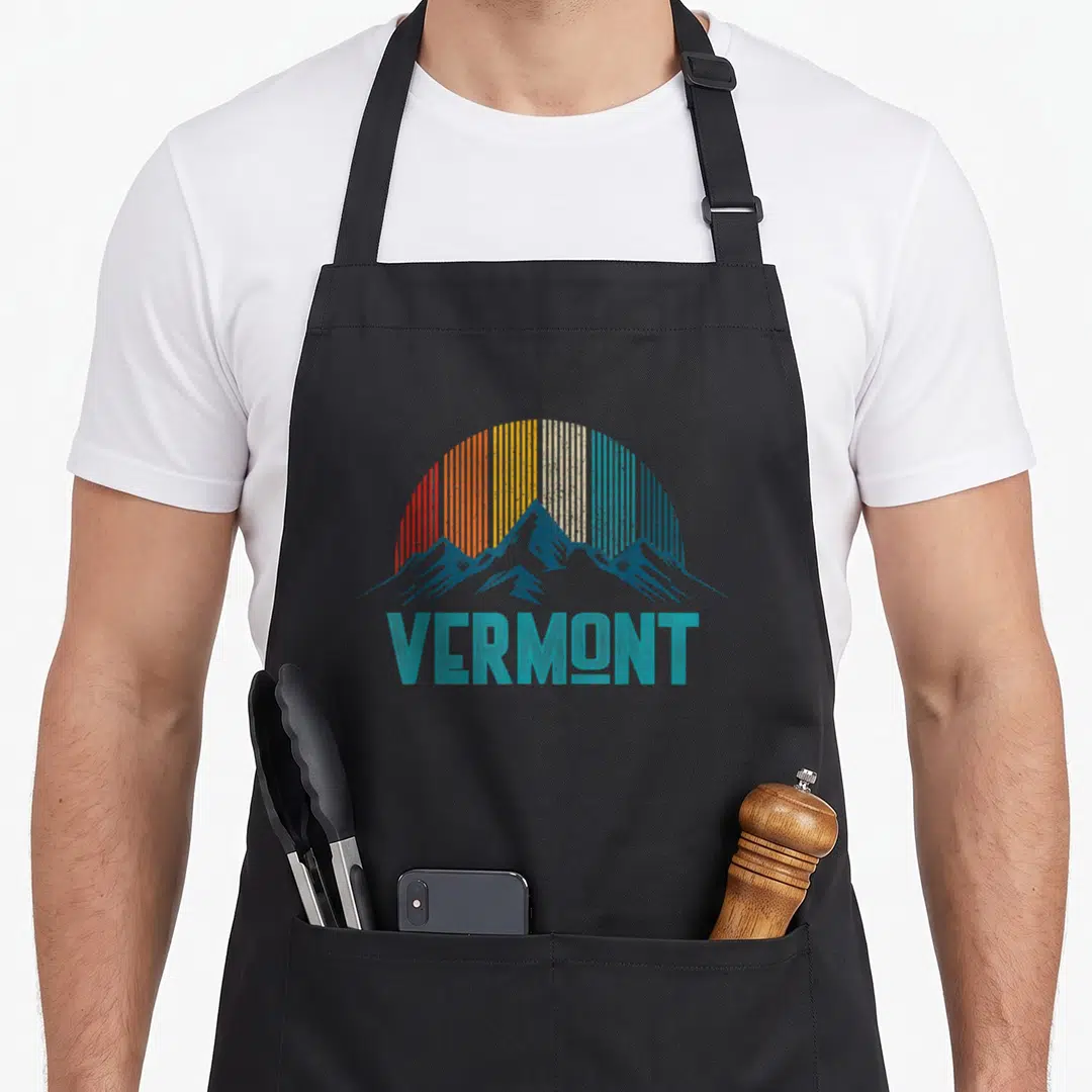 Souvenir vermont apron - Retro blue mountains under a striped sun with teal text reading VERMONT — shown on a black cotton canvas apron against a white background