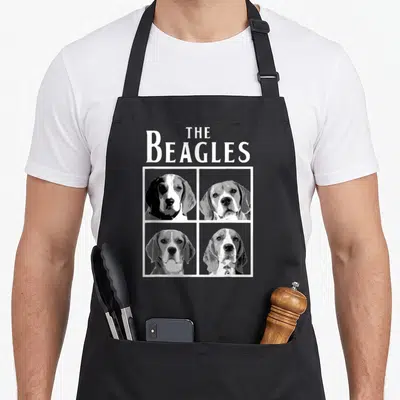 Owner beagle apron - Retro THE BEAGLES text above four beagle portraits in a white grid frame using grey, black — shown on a black cotton canvas apron against a white background