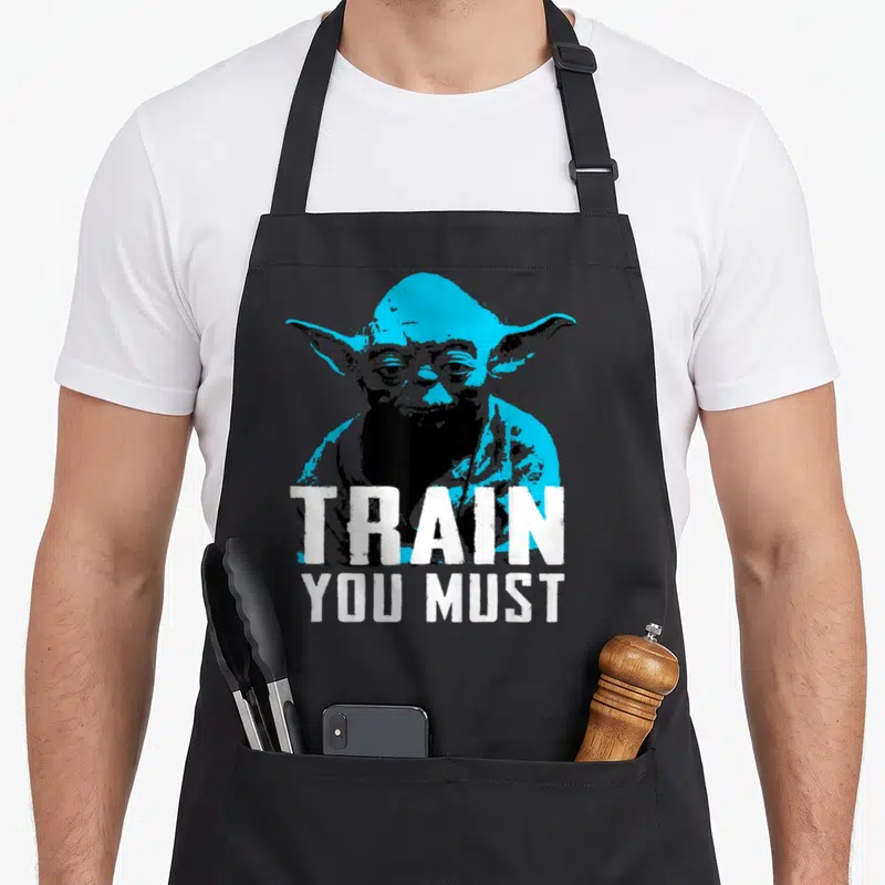Must yoda apron - Cyan and black Yoda graphic with white text reading TRAIN YOU MUST — shown on a black cotton canvas apron against a white background