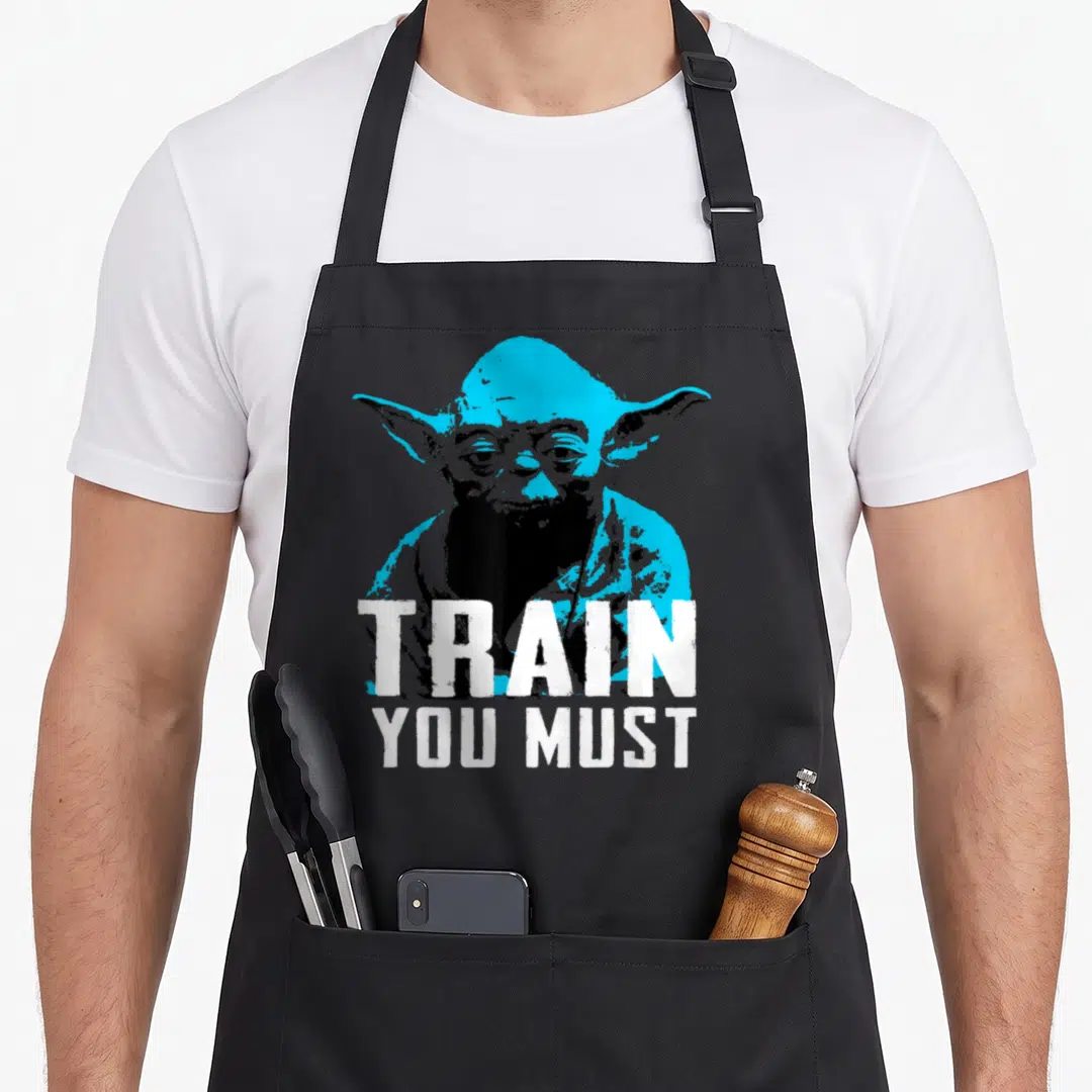 Must yoda apron - Cyan and black Yoda graphic with white text reading TRAIN YOU MUST — shown on a black cotton canvas apron against a white background