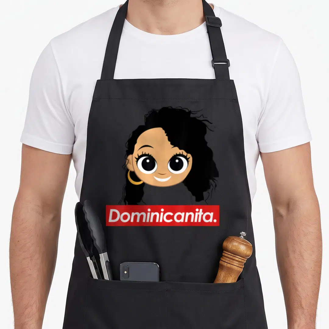 Dominican hair apron - Cartoon girl with black curly hair and gold hoop above red banner with white Dominicanita text — shown on a black cotton canvas apron against a white background