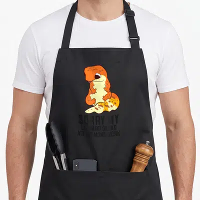 Ate leopard apron - Cartoon orange gecko on paper with text Sorry My Leopard Gecko Ate My Homework below — shown on a black cotton canvas apron against a white background
