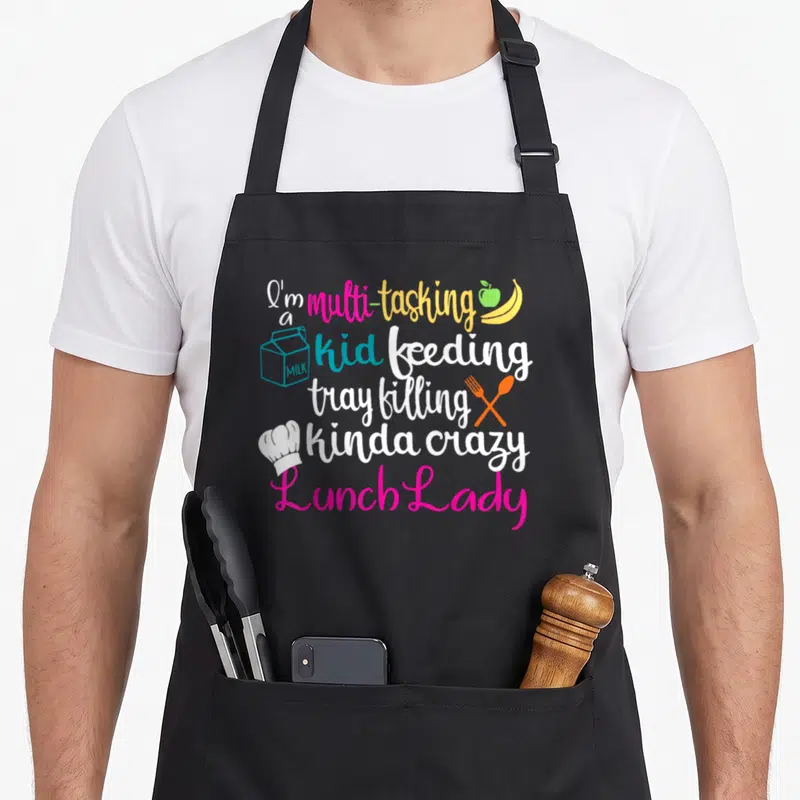 Multitasking cafeteria aprons - Stacked colorful text reading I'm a multi-tasking kid feeding tray filling kinda crazy — shown on a black cotton canvas apron against a white background