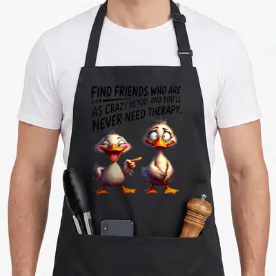 Duck find aprons - Two cartoon ducks below text about crazy friends and therapy on a black apron — shown on a black cotton canvas apron against a white background