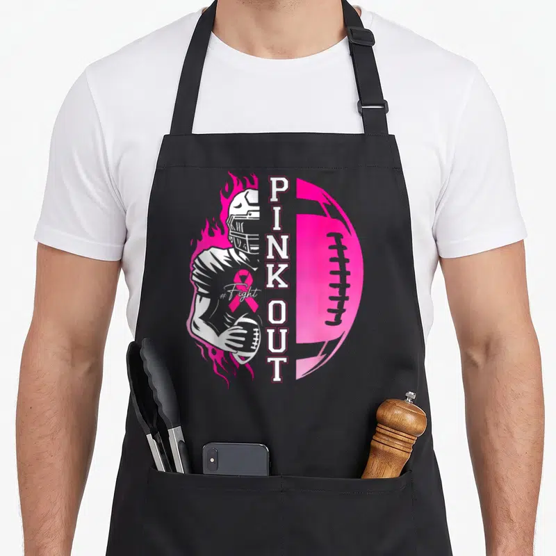 Football month breast cancer awareness aprons - Cartoon football player with PINKOUT text, pink football, and awareness — shown on a black cotton canvas apron against a white background
