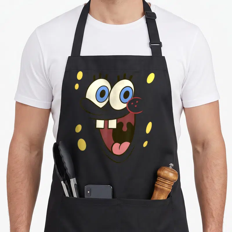 Excited face apron - SpongeBob SquarePants head with blue eyes and yellow dots scattered on grey background — shown on a black cotton canvas apron against a white background