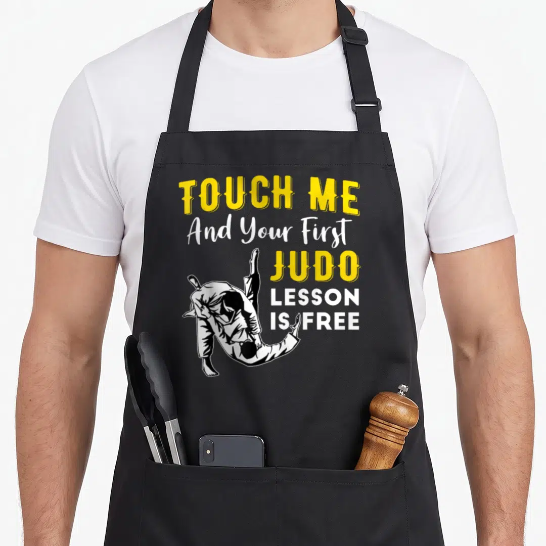 Touch judo addict apron - Cartoon judo throw illustration next to yellow and white text reading TOUCH ME And Your First — shown on a black cotton canvas apron against a white background