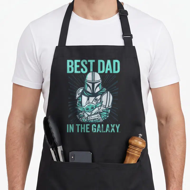Galaxy gerbil dad apron - Cartoon Mandalorian holding Grogu with mint green text BEST DAD IN THE GALAXY and grey — shown on a black cotton canvas apron against a white background