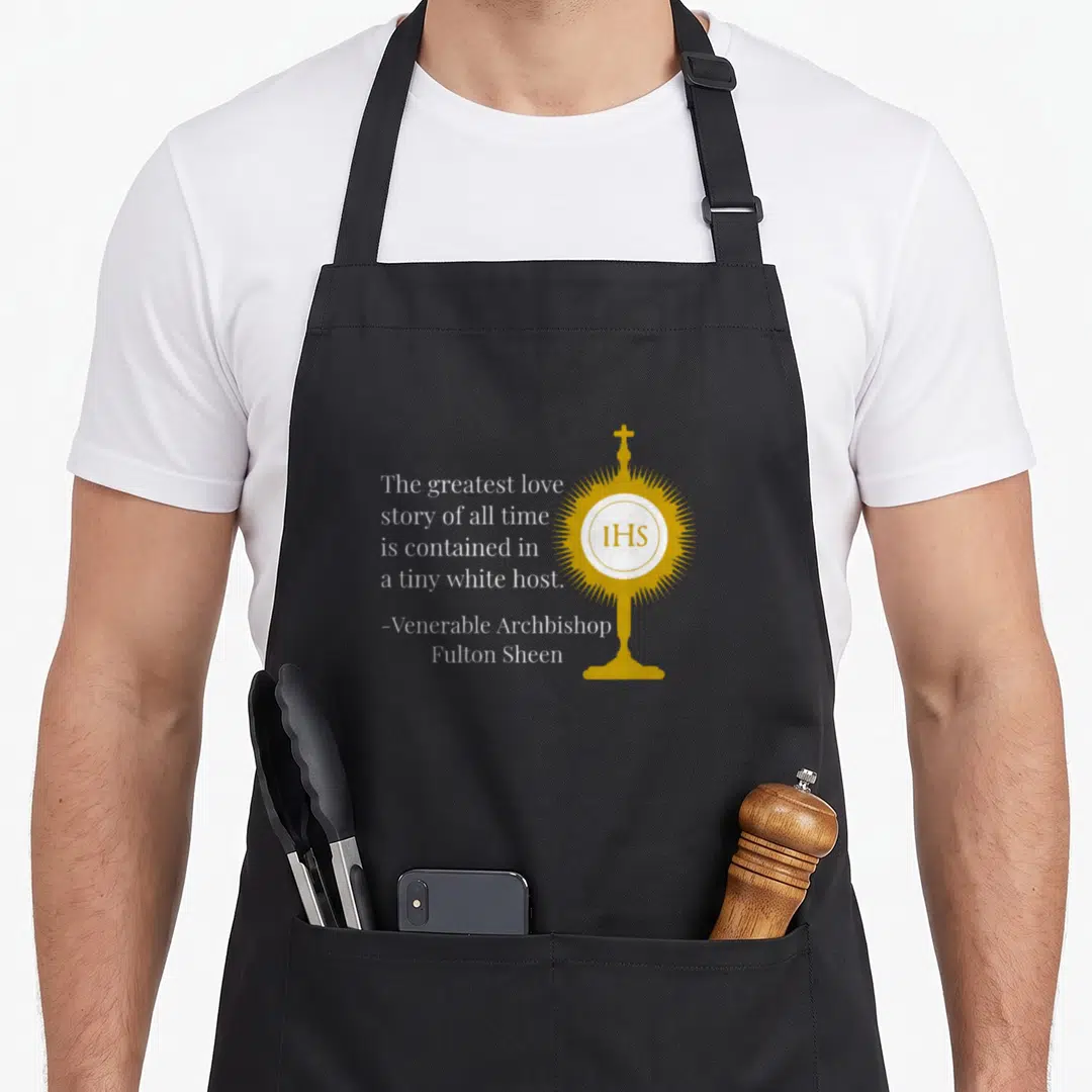 Greatest the story of the apron - Gold monstrance with IHS symbol and white text quote by Fulton Sheen on black — shown on a black cotton canvas apron against a white background