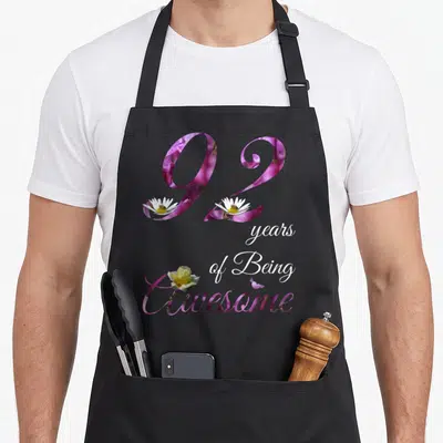 92nd floral aprons - Floral number 92 filled with purple flowers, white daisies with bees, and yellow rose text — shown on a black cotton canvas apron against a white background
