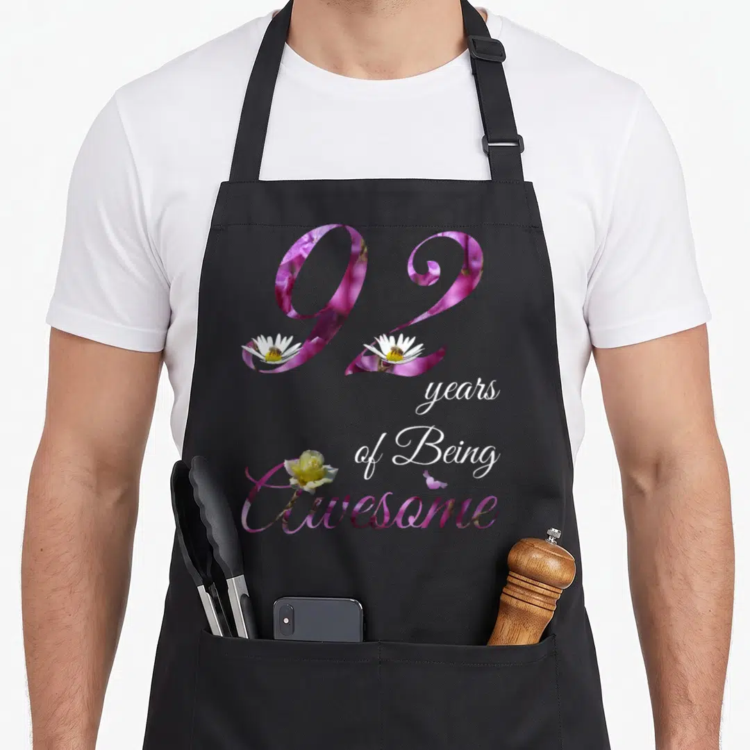 92nd floral aprons - Floral number 92 filled with purple flowers, white daisies with bees, and yellow rose text — shown on a black cotton canvas apron against a white background