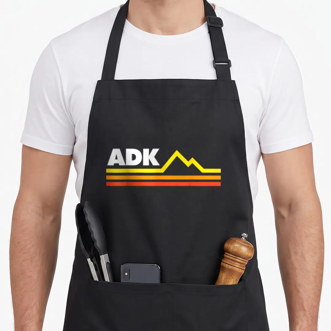 Cute adk adirondacks apron - White ADK text beside yellow mountain shape above yellow orange and red lines — shown on a black cotton canvas apron against a white background