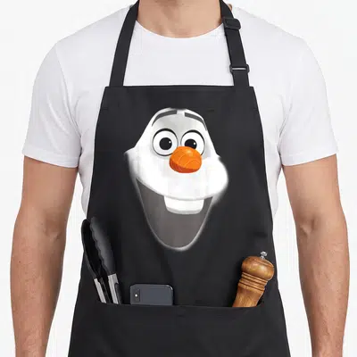 Smile snowman apron - Centered cartoon Olaf head with orange nose and big smile in white, orange, black — shown on a black cotton canvas apron against a white background