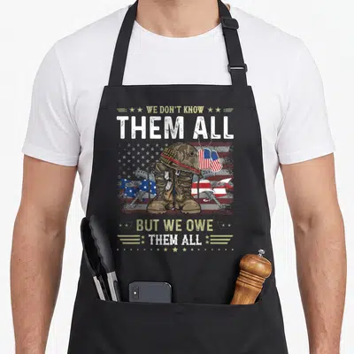 Owe veteran patriotic apron - Military helmet boots and rifles on distressed flag with We Owe Them All text — shown on a black cotton canvas apron against a white background