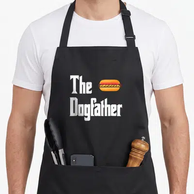 Dogfather hot dog apron - The Dogfather text with a hot dog graphic in orange, brown, and yellow — shown on a black cotton canvas apron against a white background