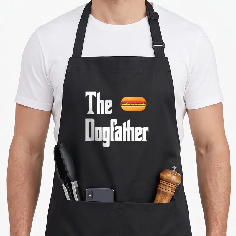 Dogfather hot dog apron - The Dogfather text with a hot dog graphic in orange, brown, and yellow — shown on a black cotton canvas apron against a white background