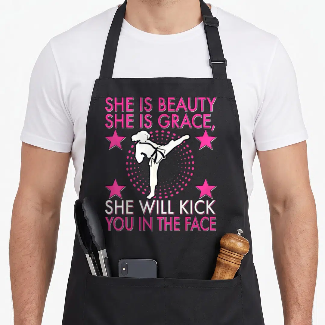 Taekwondo kick karate apron - Pink and white cartoon martial artist kicking surrounded by stars and She Is Beauty She — shown on a black cotton canvas apron against a white background