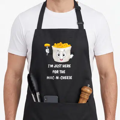 Funny cheese mac apron - Cartoon mac and cheese bowl with fork and I'M JUST HERE FOR THE MAC-N-CHEESE text — shown on a black cotton canvas apron against a white background