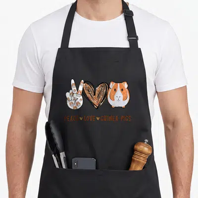 Peace guinea lover apron - Cartoon hand peace sign, gold heart, and guinea pig above text PEACE LOVE GUINEA PIGS — shown on a black cotton canvas apron against a white background
