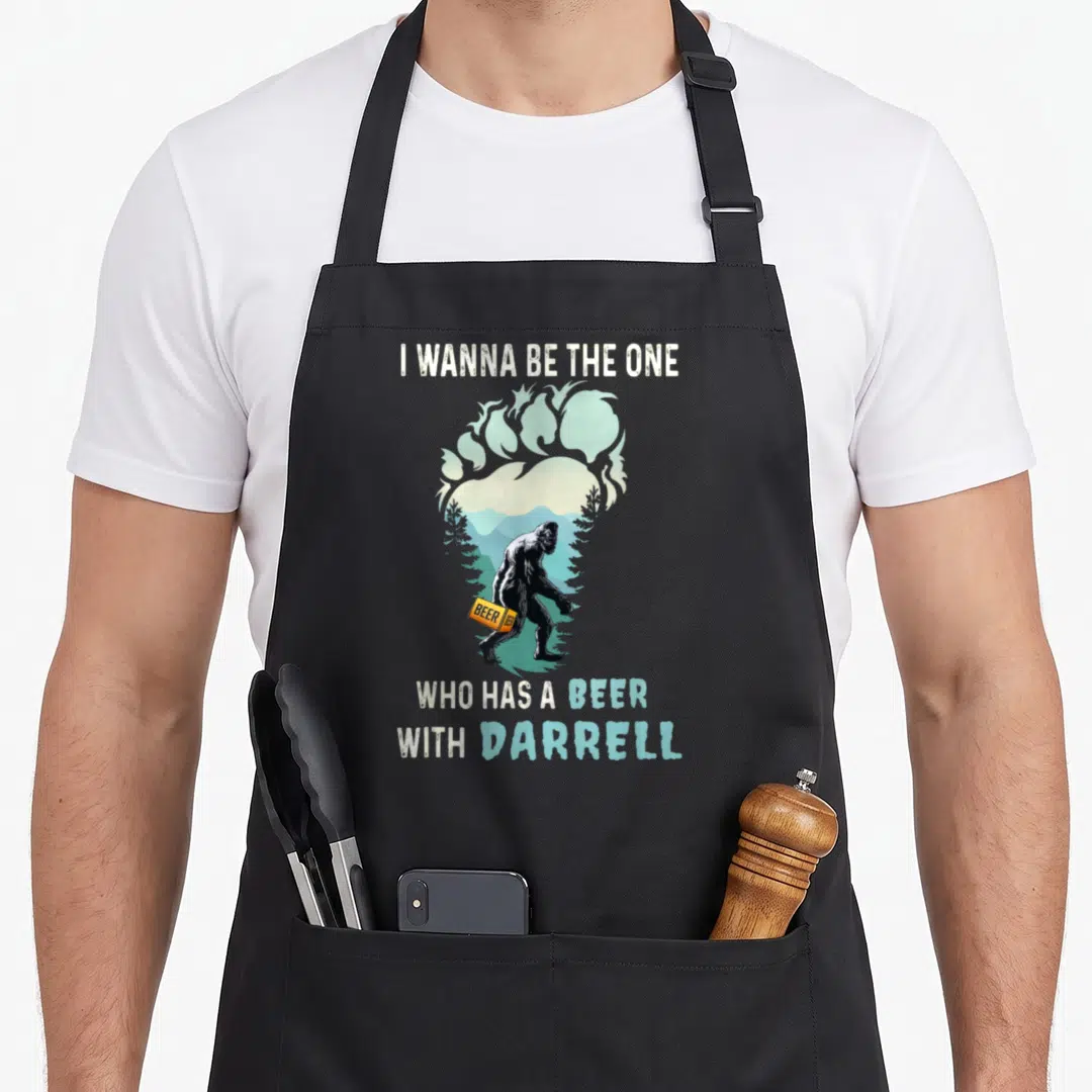Darrell beer apron - Bigfoot holding orange beer can inside footprint mountain landscape with white and blue text — shown on a black cotton canvas apron against a white background