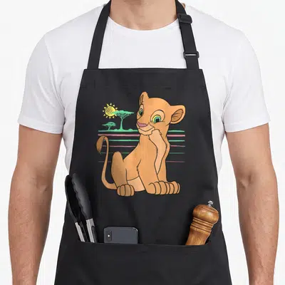 Nala lion apron - Cartoon lion cub sitting with teal trees and pink horizontal lines on black apron — shown on a black cotton canvas apron against a white background