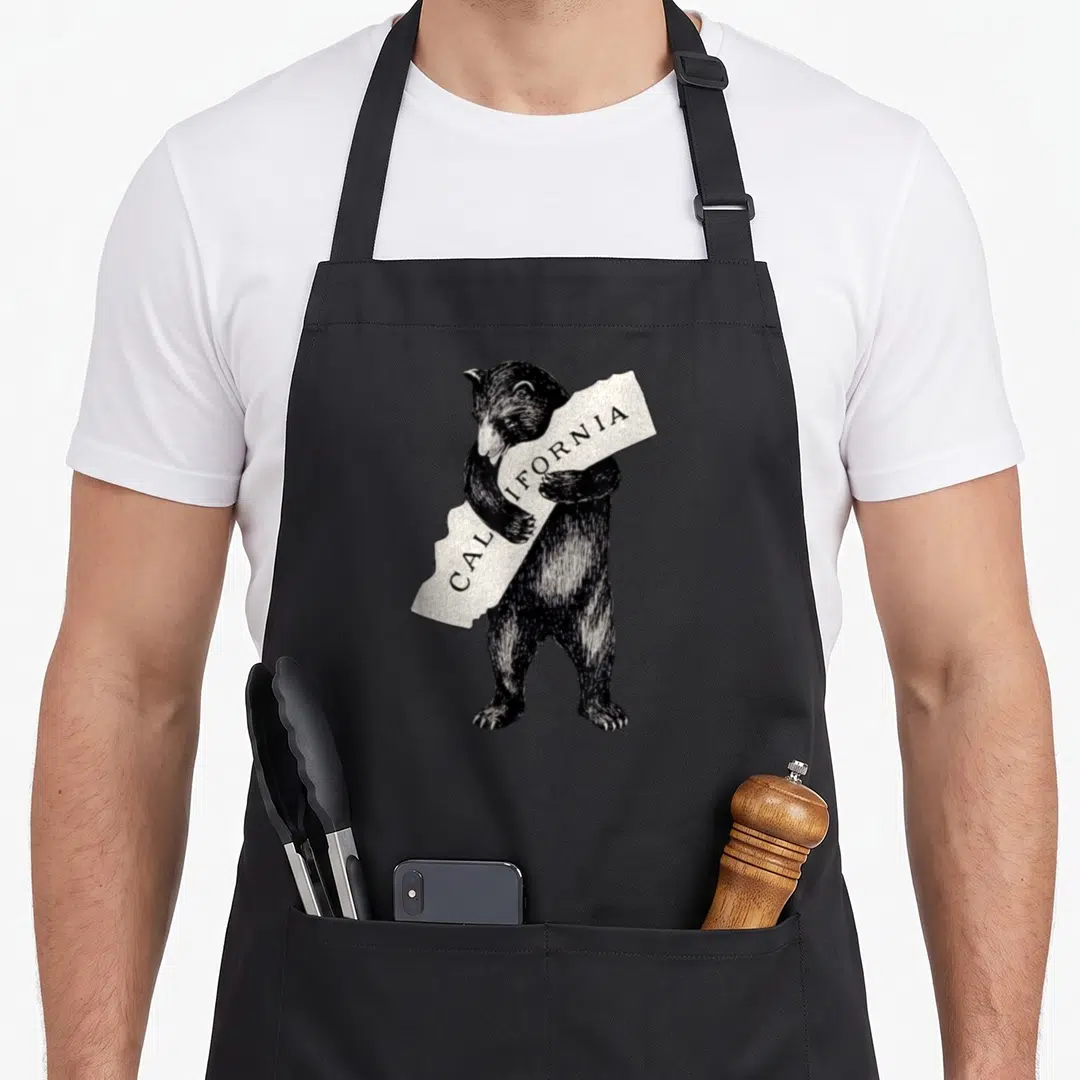 Pride bear apron - Black bear standing holding white banner with CALIFORNIA text — shown on a black cotton canvas apron against a white background