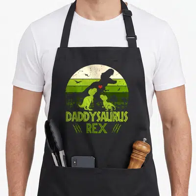Daddysaurus father day apron - Retro T-Rex silhouette over two baby dinosaurs with DADDYSAURUS REX text and red heart — shown on a black cotton canvas apron against a white background