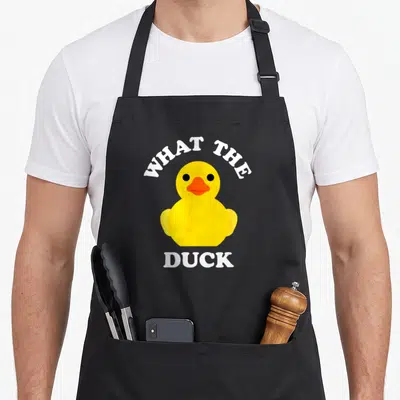 Ducky yellow rubber apron - Cartoon yellow rubber duck with orange beak and white text reading What The Duck — shown on a black cotton canvas apron against a white background