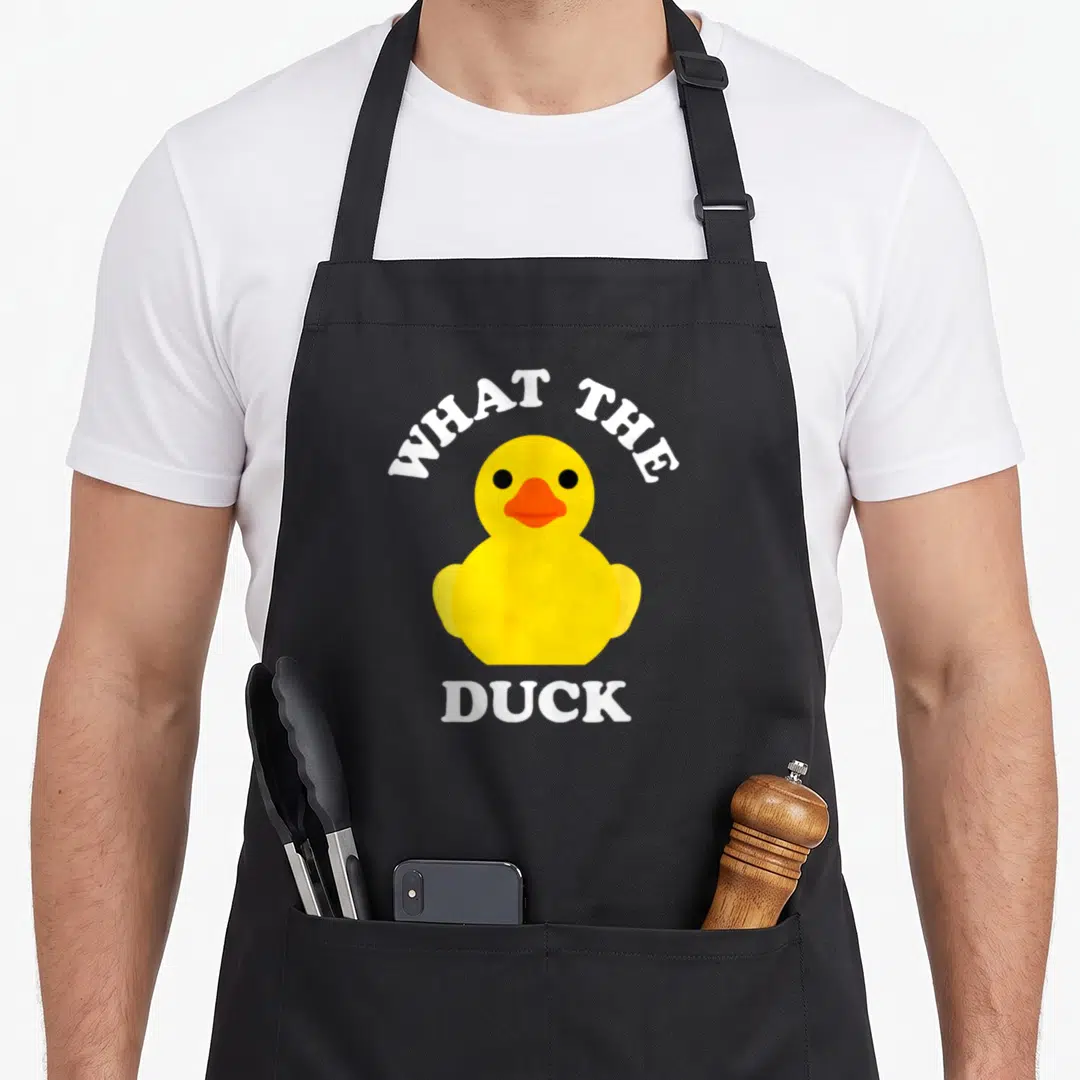 Ducky yellow rubber apron - Cartoon yellow rubber duck with orange beak and white text reading What The Duck — shown on a black cotton canvas apron against a white background