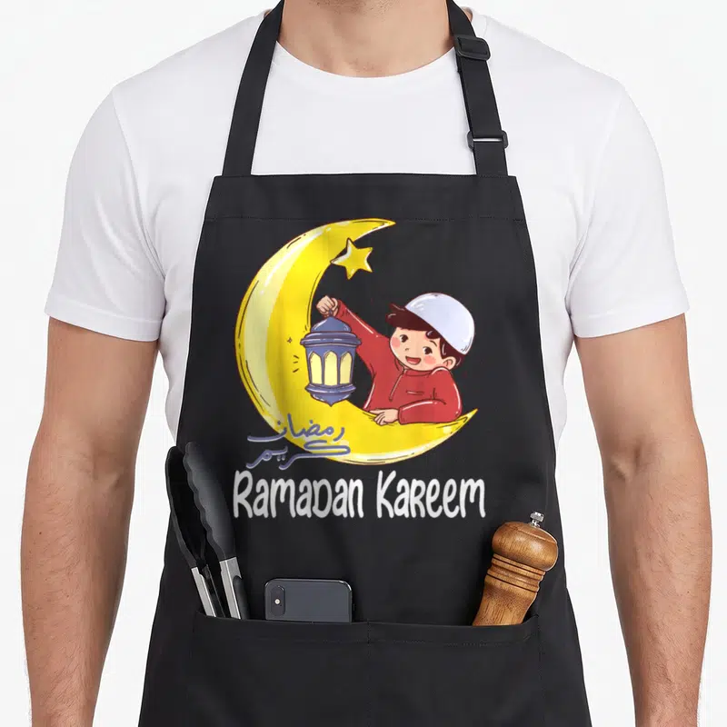 Ramadan arab boy apron - Cartoon boy in yellow crescent moon holding blue lantern with Ramadan Kareem text — shown on a black cotton canvas apron against a white background