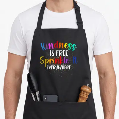 Free sprinkle apron - Stacked text Kindness IS FREE Sprinkle It EVERYWHERE with multicolored dots on black fabric — shown on a black cotton canvas apron against a white background