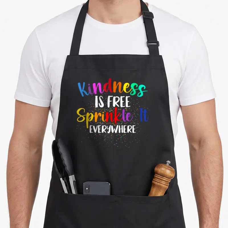 Free sprinkle apron - Stacked text Kindness IS FREE Sprinkle It EVERYWHERE with multicolored dots on black fabric — shown on a black cotton canvas apron against a white background