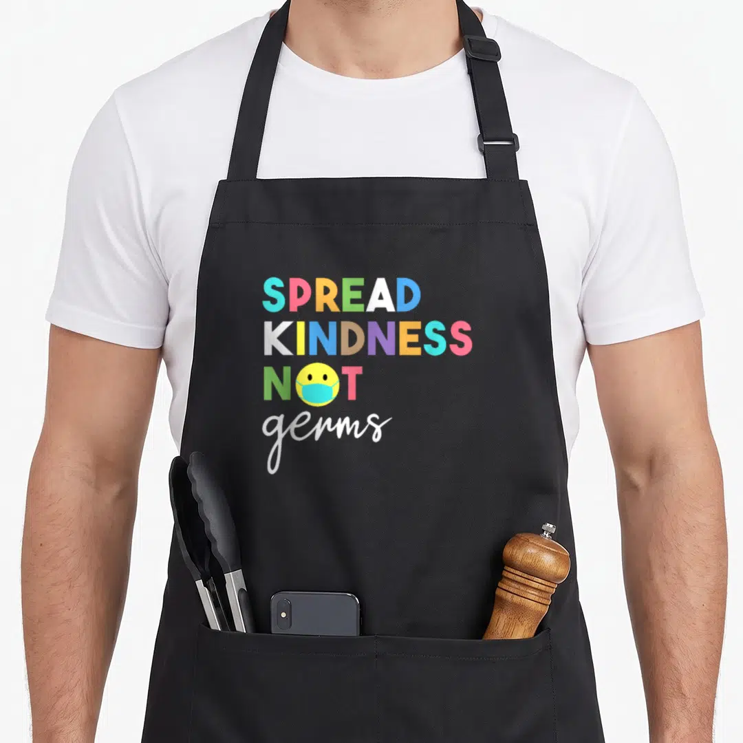 Spread essential apron - Stacked colorful text reading SPREAD KINDNESS NOT germs with a yellow smiley face wearing — shown on a black cotton canvas apron against a white background