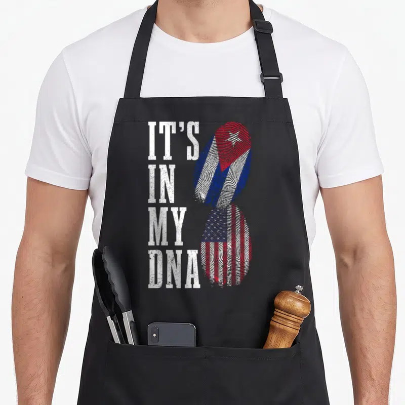 Cuba dna american flag apron - IT'S IN MY DNA text beside two fingerprint shapes with Cuban and American flag patterns — shown on a black cotton canvas apron against a white background