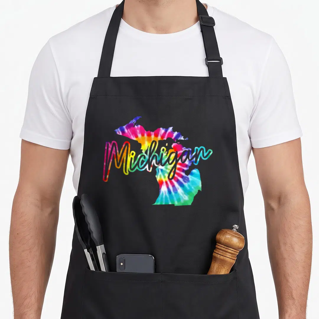 Michigan tie-dye apron - Tie-dye map of Michigan in blue pink yellow green and red with script text overlay — shown on a black cotton canvas apron against a white background