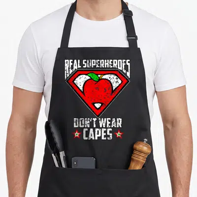 Distressed teacher apron - Red apple inside shield with text Real Superheroes Don't Wear Capes — shown on a black cotton canvas apron against a white background