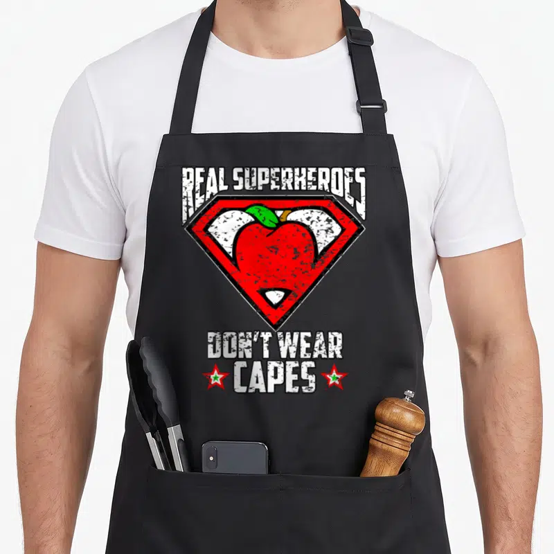 Distressed teacher apron - Red apple inside shield with text Real Superheroes Don't Wear Capes — shown on a black cotton canvas apron against a white background