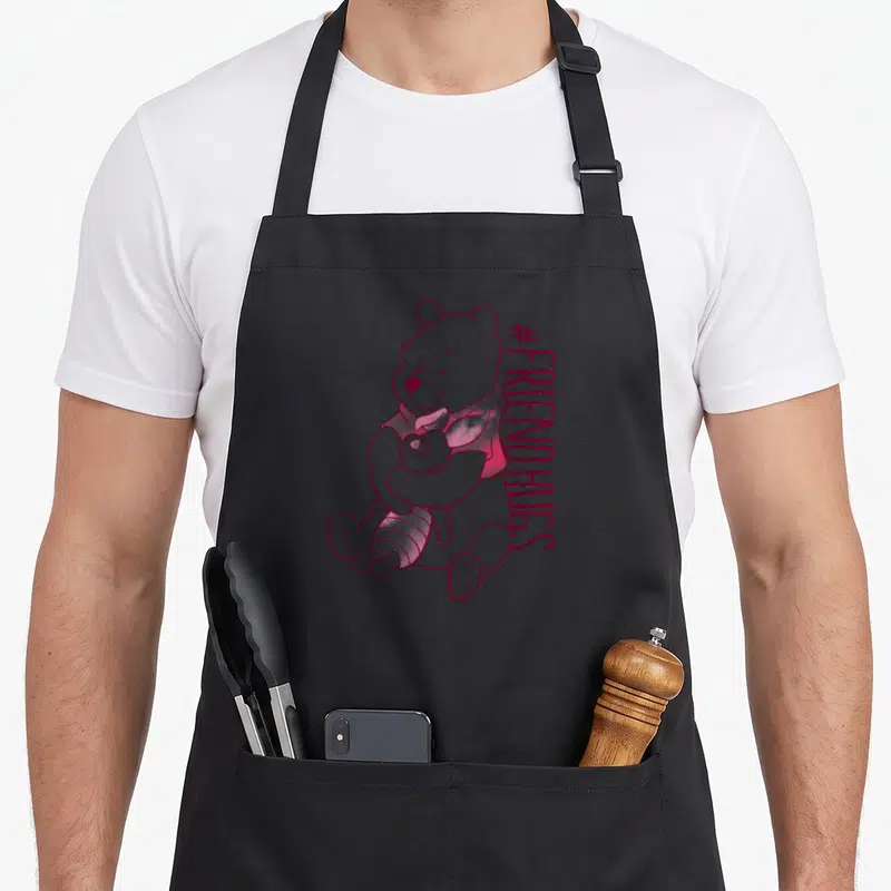 Hug friends apron - Winnie the Pooh hugging Piglet in dark pink line-art beside vertical #FRIENDHUGS text — shown on a black cotton canvas apron against a white background