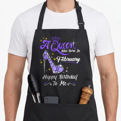 February birthday baking queen apron - Purple high heel shoe with A Queen Was Born In February text and yellow stars — shown on a black cotton canvas apron against a white background
