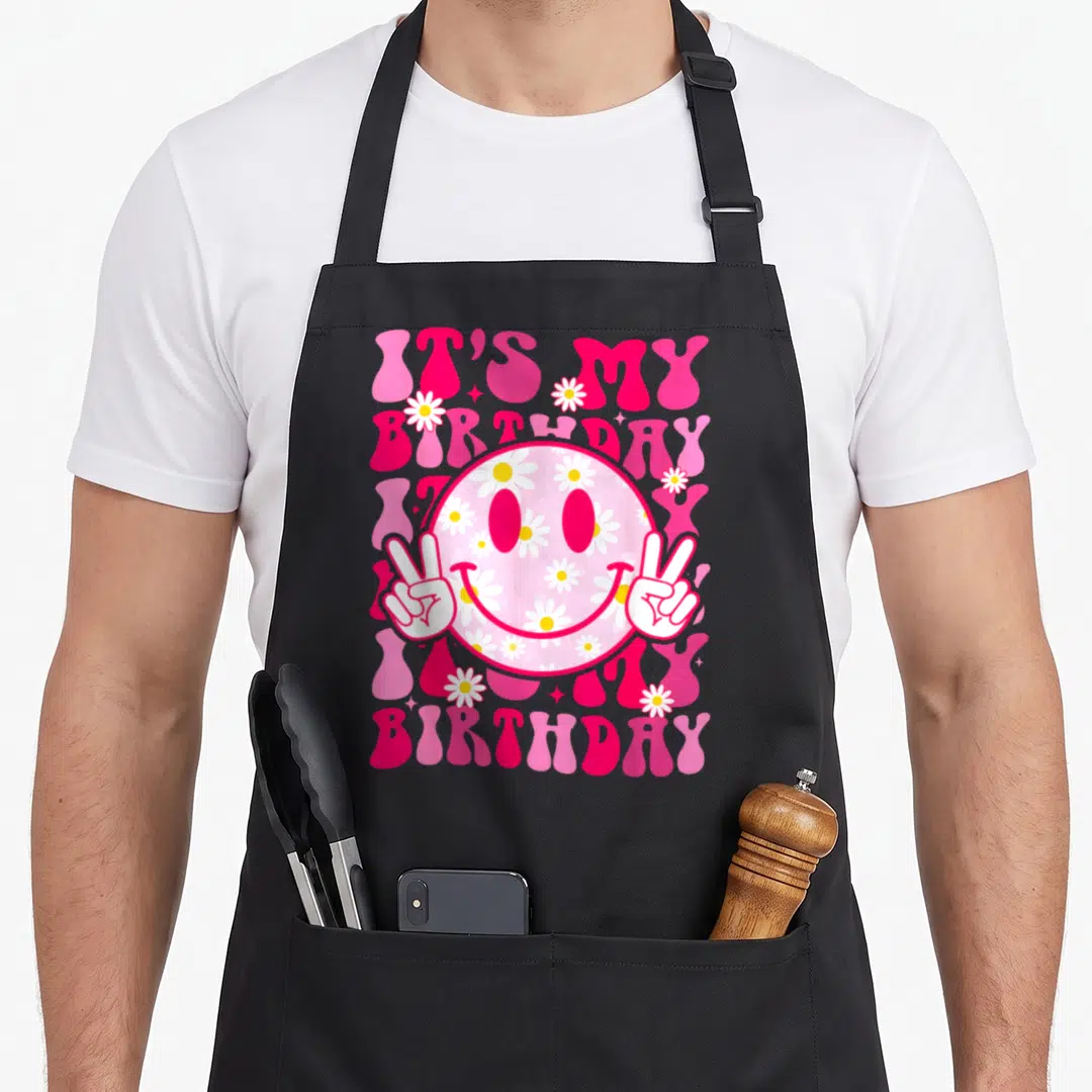 Preppy custom face apron - Retro pink smiley face with daisies, peace hands, and IT'S MY BIRTHDAY text — shown on a black cotton canvas apron against a white background