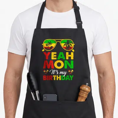 Yeah birthday apron - Retro sunglasses with rasta colors and YEAH MON It's my BIRTHDAY text flanked by maracas — shown on a black cotton canvas apron against a white background