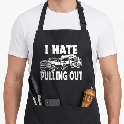 Travel camping trailer apron - White line art of truck towing camper with I HATE PULLING OUT text — shown on a black cotton canvas apron against a white background