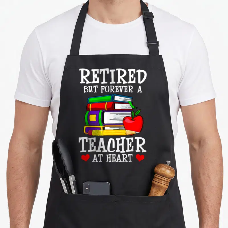 But teachers apron - Cartoon stack of books, red apple, pencil, and text Retired But Forever a Teacher at Heart — shown on a black cotton canvas apron against a white background