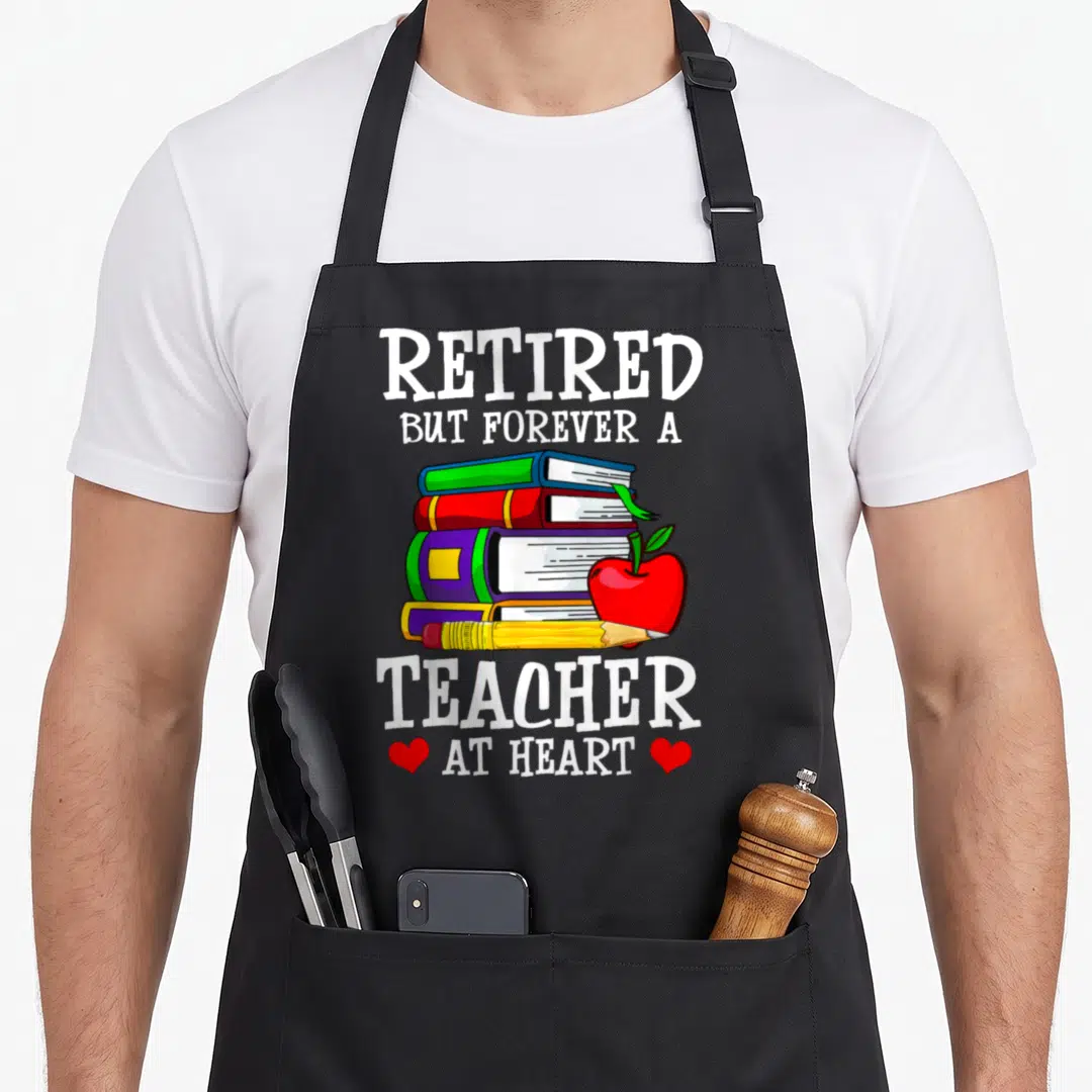 But teachers apron - Cartoon stack of books, red apple, pencil, and text Retired But Forever a Teacher at Heart — shown on a black cotton canvas apron against a white background