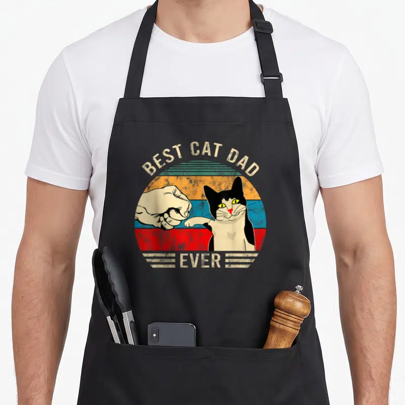 Funny bump cat apron - Best Cat Dad Ever text over human and tuxedo cat fist bump in vintage striped circle — shown on a black cotton canvas apron against a white background